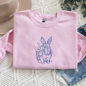 May include: A pink sweatshirt with a blue embroidered image of a rabbit with a bow. The rabbit is facing forward and has its ears up.