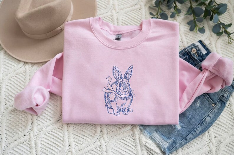 May include: A pink sweatshirt with a blue embroidered image of a rabbit with a bow. The rabbit is facing forward and has its ears up.