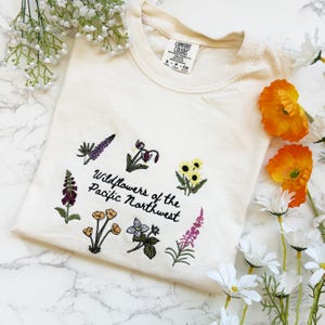Pode incluir: Camisola creme com bordados de flores silvestres e o texto "Wildflowers of the Pacific Northwest". O design apresenta v&aacute;rias ilustra&ccedil;&otilde;es florais coloridas. A camisola tem uma etiqueta que diz "Comfort Colors".