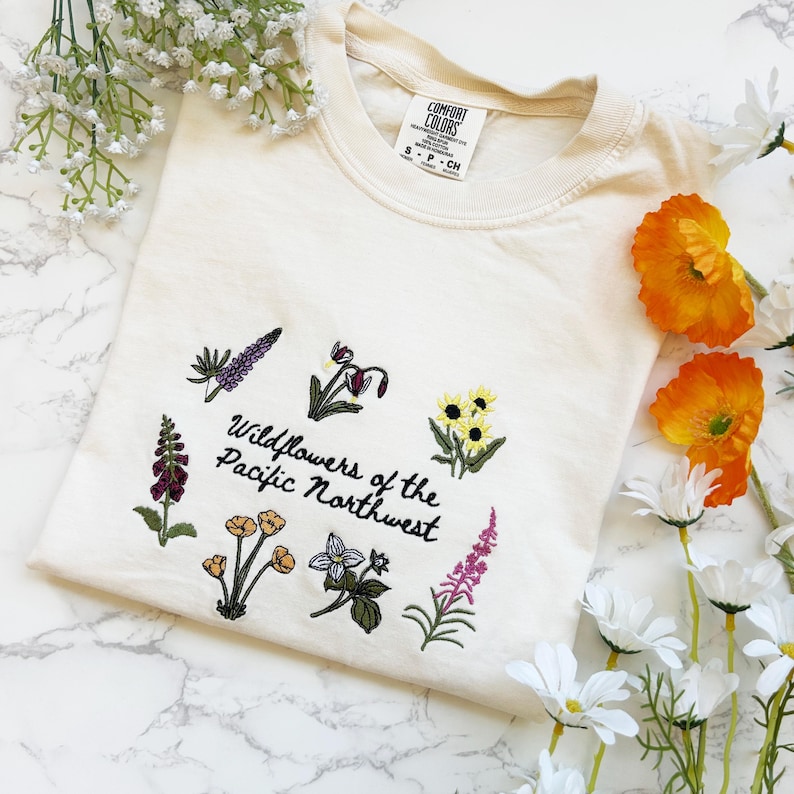 Pode incluir: Camisola creme com bordados de flores silvestres e o texto "Wildflowers of the Pacific Northwest". O design apresenta v&aacute;rias ilustra&ccedil;&otilde;es florais coloridas. A camisola tem uma etiqueta que diz "Comfort Colors".
