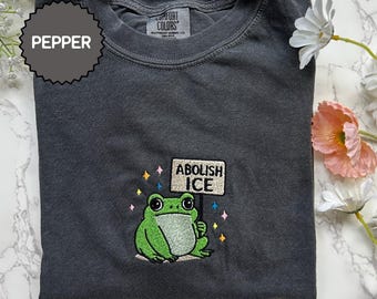 May include: Pepper-colored crew neck sweatshirt with an embroidered green frog holding a sign that reads "ABOLISH ICE." The design includes colorful stars. The sweatshirt is from Comfort Colors.