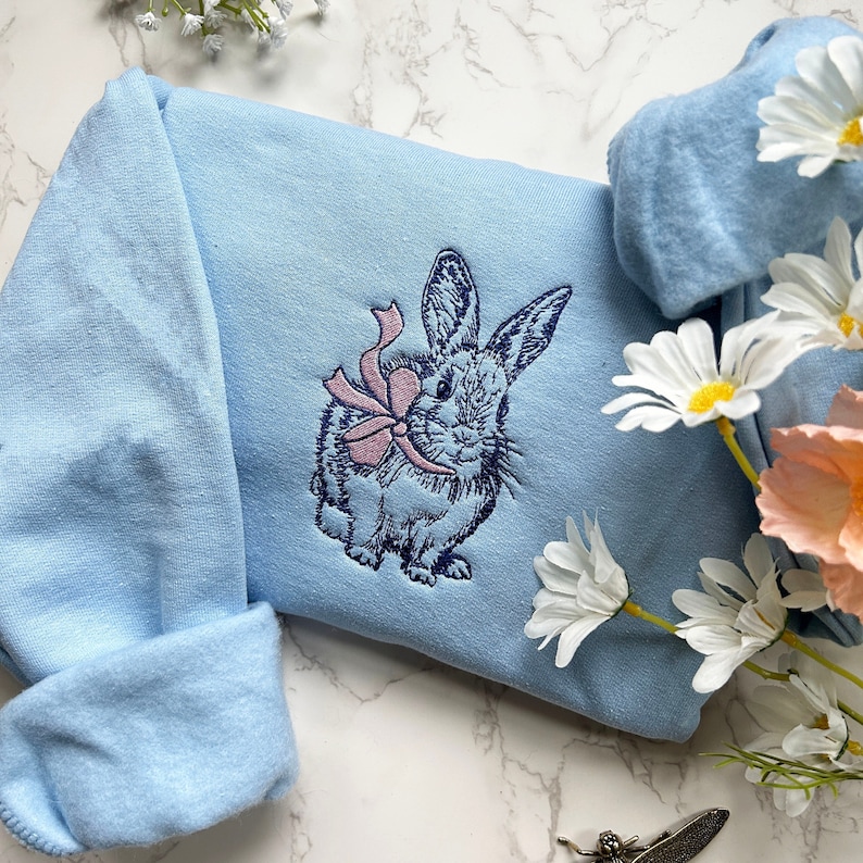 May include: A light blue sweatshirt with an embroidered design of a rabbit holding a pink bow. The rabbit is in a light blue color with dark blue outlines.