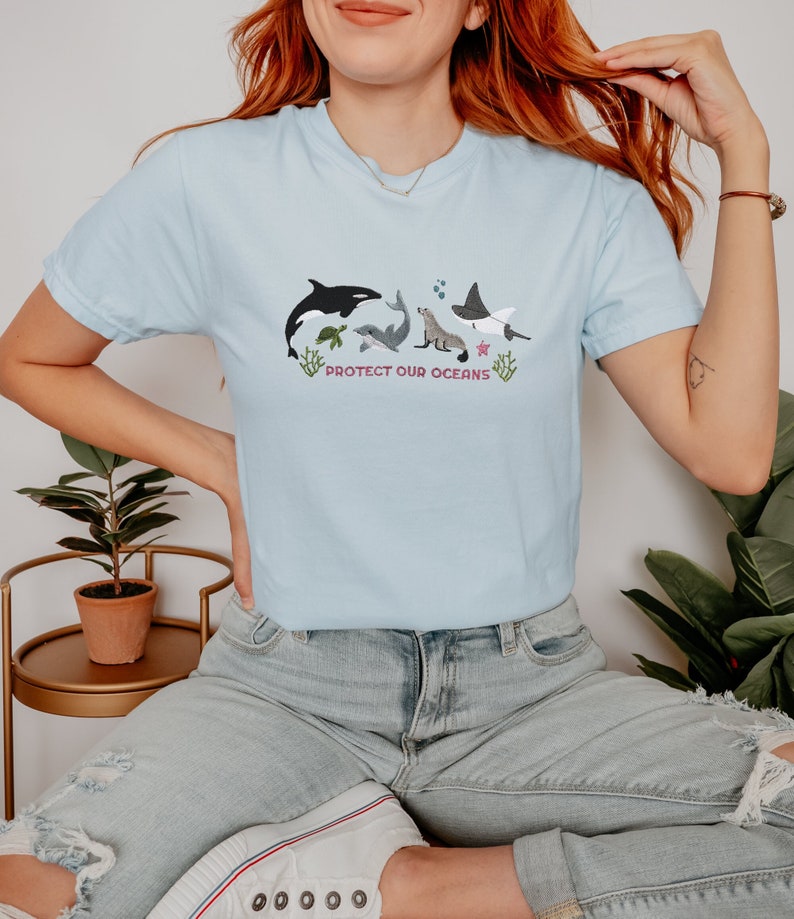 May include: A light blue t-shirt with an embroidered design of various sea creatures, including an orca, a sea turtle, a dolphin, a seal, and a manta ray. The design also includes coral and a starfish. The text "PROTECT OUR OCEANS" is embroidered below the sea creatures.