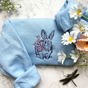 May include: A light blue sweatshirt with an embroidered image of a rabbit wearing a pink bow. The sweatshirt is on a white marble surface.