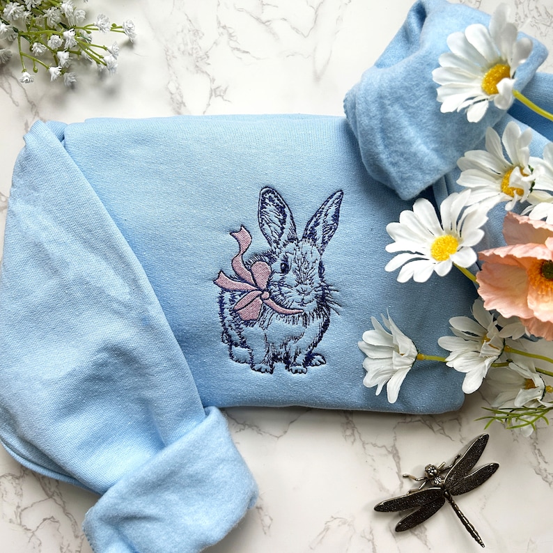 May include: A light blue sweatshirt with an embroidered image of a rabbit wearing a pink bow. The sweatshirt is on a white marble surface.