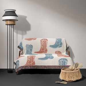 May include: A white throw blanket with a fringe border features a repeated pattern of blue and rust-colored cowboy boots. The blanket is draped over a small sofa with a black metal frame. A black floor lamp with a white and black lampshade is in the background.