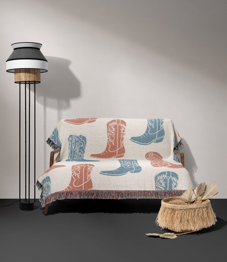 May include: A white throw blanket with a fringe border features a repeated pattern of blue and rust-colored cowboy boots. The blanket is draped over a small sofa with a black metal frame. A black floor lamp with a white and black lampshade is in the background.