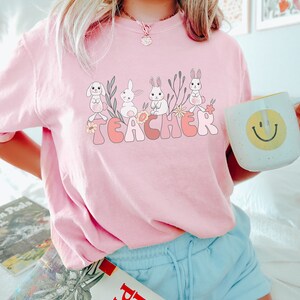 Op de afbeelding: Roze T-shirt met een afbeelding van konijntjes en bloemen die "TEACHER" spellen.