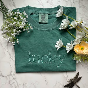 May include: A green t-shirt with the word "lucky" embroidered in white with green leaves. The shirt is made by Comfort Colors and has a tag on the front.