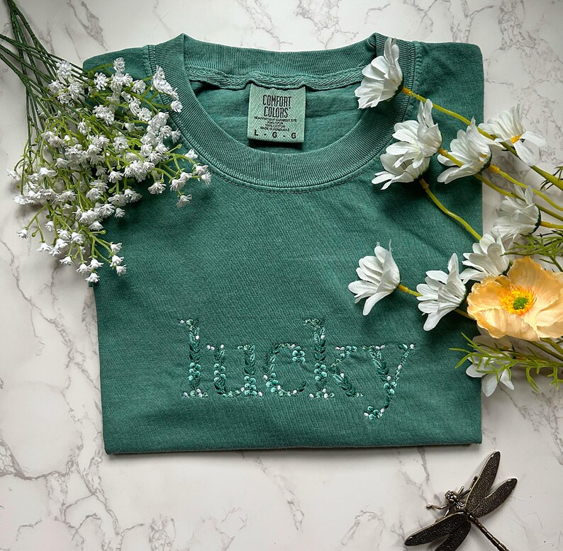 May include: A green t-shirt with the word "lucky" embroidered in white with green leaves. The shirt is made by Comfort Colors and has a tag on the front.