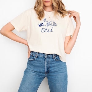 May include: Cream-colored t-shirt with a navy blue graphic of a hand holding a croissant and the word "oui" underneath. The model is wearing blue jeans. The shirt is a casual style.