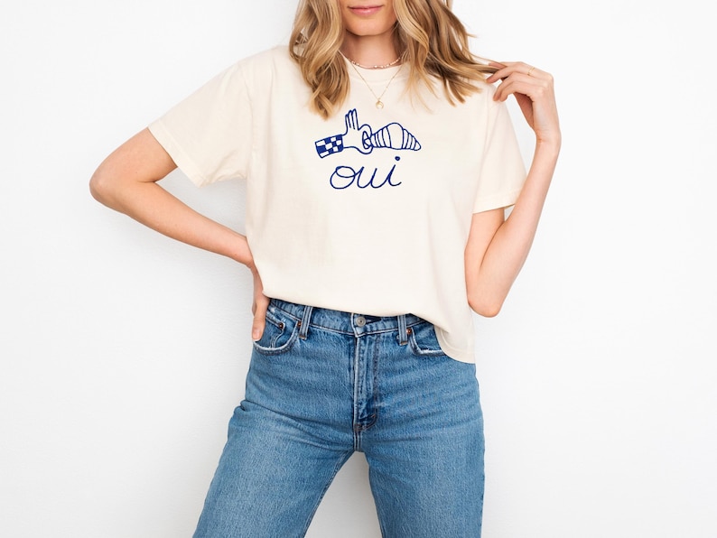 May include: Cream-colored t-shirt with a navy blue graphic of a hand holding a croissant and the word "oui" underneath. The model is wearing blue jeans. The shirt is a casual style.