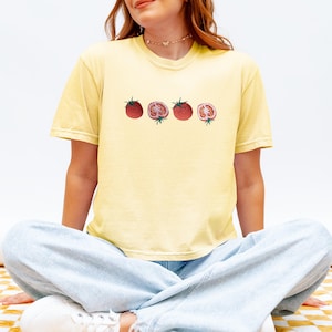 May include: A light yellow t-shirt featuring a watercolor illustration of four tomatoes. The tomatoes are red with green stems, two whole and two sliced open, showcasing the inside. The shirt is worn by a person sitting cross-legged.