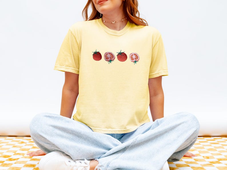 May include: A light yellow t-shirt featuring a watercolor illustration of four tomatoes. The tomatoes are red with green stems, two whole and two sliced open, showcasing the inside. The shirt is worn by a person sitting cross-legged.