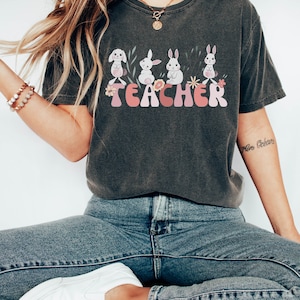 Op de afbeelding: Een donkergrijs T-shirt met een afbeelding van vier witte konijnen en het woord "TEACHER" in roze en witte letters. De konijnen zijn omringd door groene bladeren en roze bloemen.