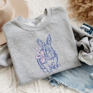 May include: A gray sweatshirt with a blue and pink embroidered design of a rabbit holding a bow.