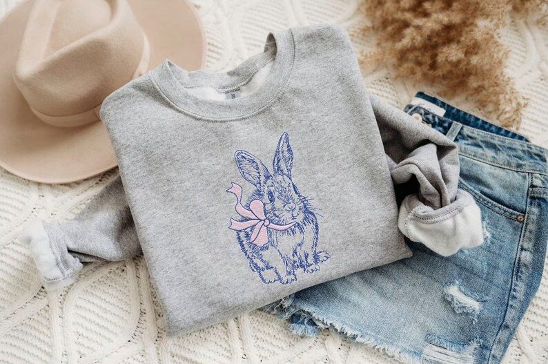 May include: A gray sweatshirt with a blue and pink embroidered design of a rabbit holding a bow.
