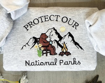 May include: Gray sweatshirt with embroidered text "Protect Our National Parks" featuring a brown bear sitting in a chair by a campfire in a mountain landscape.