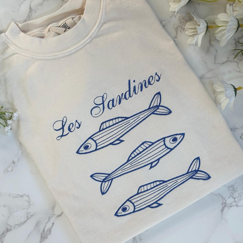 May include: A white t-shirt with blue embroidery of three sardines and the text "Les Sardines".