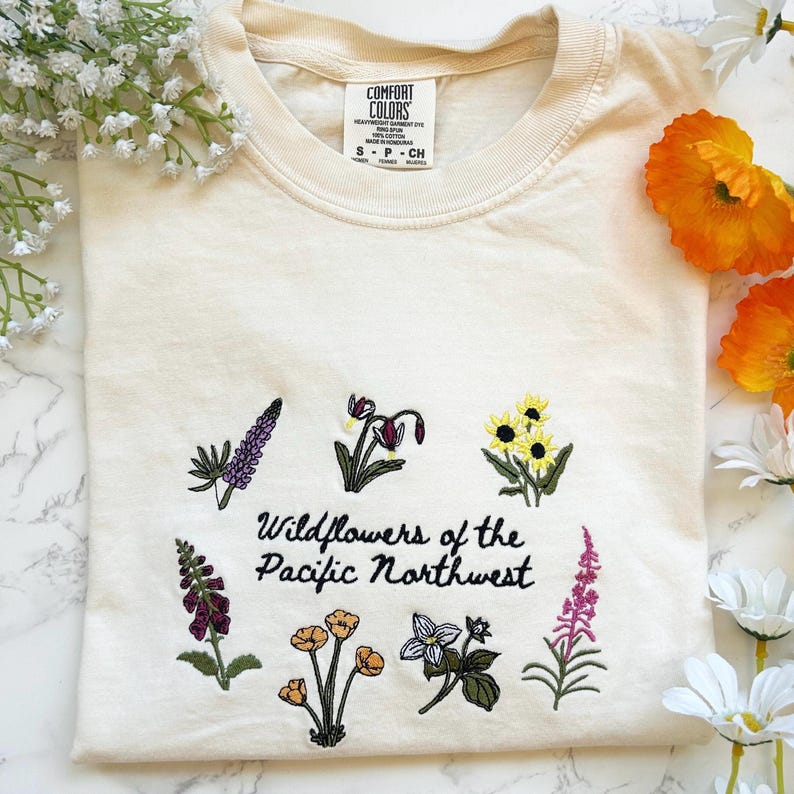 Pode incluir: Camiseta creme com bordados de flores silvestres e o texto "Wildflowers of the Pacific Northwest". O design apresenta v&aacute;rias ilustra&ccedil;&otilde;es florais coloridas. A camiseta tem gola redonda e mangas curtas.
