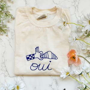May include: Cream-colored sweatshirt with a blue embroidered design of a hand holding a croissant, and the word "oui" below. The sweatshirt is surrounded by white daisies and a single orange flower.