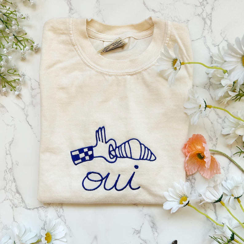 May include: Cream-colored sweatshirt with a blue embroidered design of a hand holding a croissant, and the word "oui" below. The sweatshirt is surrounded by white daisies and a single orange flower.