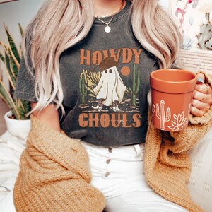 K&ouml;nnte beinhalten: Ein dunkelgraues T-Shirt mit einem Aufdruck eines Geistes, der einen Cowboyhut und Stiefel tr&auml;gt. Der Text "Howdy Ghouls" ist auf dem Shirt gedruckt.