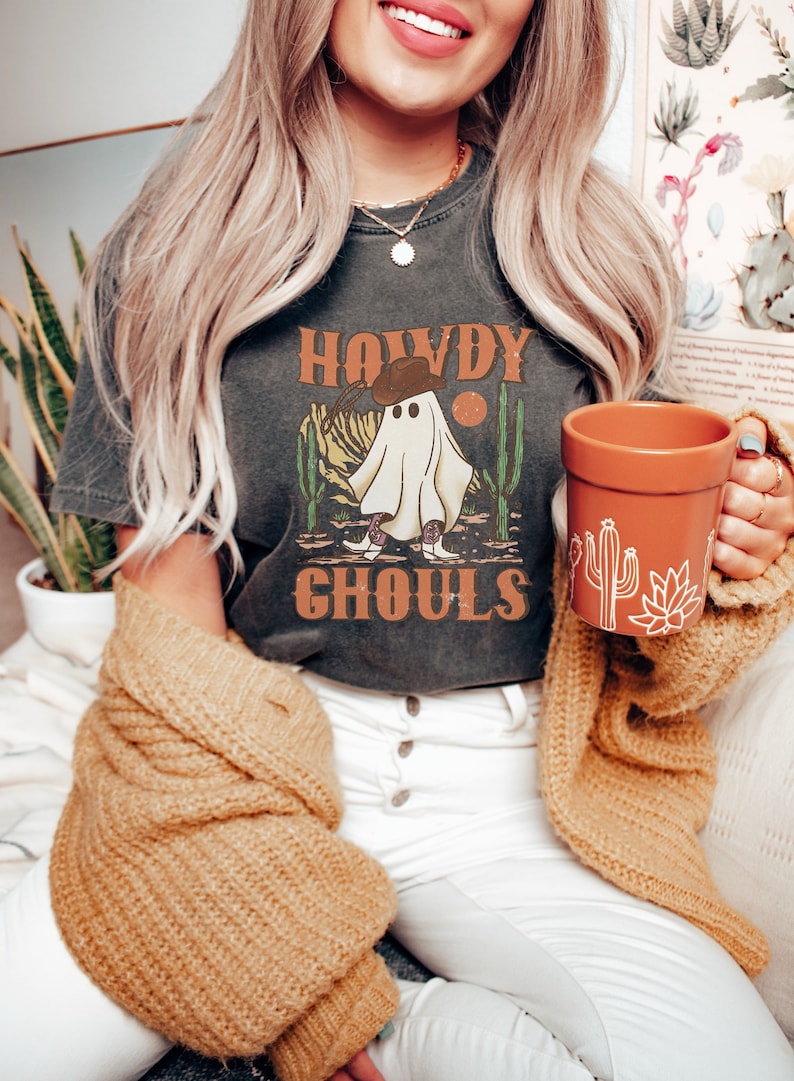 K&ouml;nnte beinhalten: Ein dunkelgraues T-Shirt mit einem Aufdruck eines Geistes, der einen Cowboyhut und Stiefel tr&auml;gt. Der Text "Howdy Ghouls" ist auf dem Shirt gedruckt.