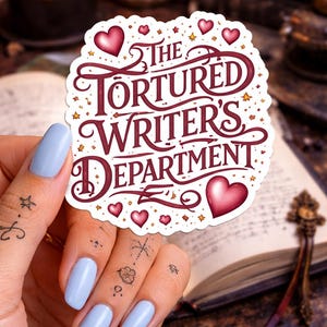 May include: A round sticker with the text "The Tortured Writer's Department" in burgundy script, adorned with hearts and stars. The sticker is held up by a hand with light blue nails, with a blurred background of a book and candle.