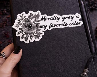 May include: A black journal with a sticker that reads "Morally gray is my favorite color." The sticker features a floral design in black and white. A black pen rests on the journal, and a skull charm hangs from the bookmark.