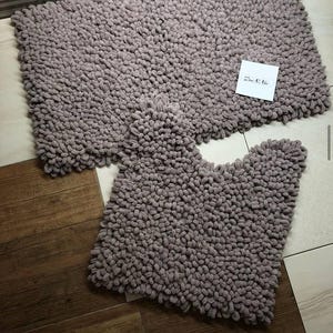 May include: A mauve-coloured two-piece bathroom rug set. The set comprises a rectangular bath mat and a smaller, contoured rug. Both rugs have a thick, textured, looped pile. A small white tag with text is visible.