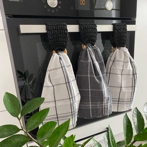 May include: Three kitchen towels with black crochet tops and wooden rings hanging on a stainless steel bar. The towels are in a plaid pattern, with colors of gray and white. A green plant with large leaves is in the foreground.