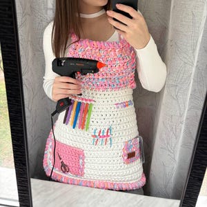 May include: A handmade crochet apron in white, pink, and pastel colors. The apron features a bib with a pink and pastel top, a white body, and pockets. It has a colorful trim and holds a glue gun and craft supplies.