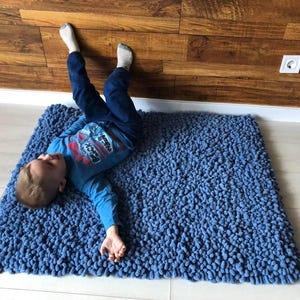 Peut inclure: Un tapis bleu touffu avec un enfant allong&eacute; dessus. Le tapis est rectangulaire et a une surface textur&eacute;e. L'enfant porte une chemise bleue &agrave; manches longues et un pantalon bleu.