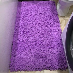Peut inclure: Un tapis de bain rectangulaire et moelleux de couleur violette. Le tapis est fait d'une mati&egrave;re &eacute;paisse et textur&eacute;e et est plac&eacute; sur un sol carrel&eacute;. Le tapis est positionn&eacute; devant des toilettes blanches et une machine &agrave; laver.