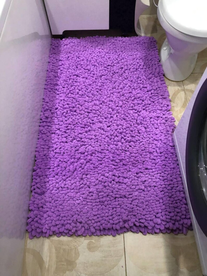 Peut inclure: Un tapis de bain rectangulaire et moelleux de couleur violette. Le tapis est fait d'une mati&egrave;re &eacute;paisse et textur&eacute;e et est plac&eacute; sur un sol carrel&eacute;. Le tapis est positionn&eacute; devant des toilettes blanches et une machine &agrave; laver.