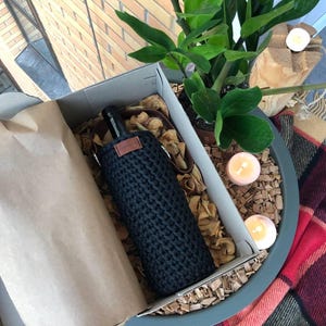 K&ouml;nnte beinhalten: Eine schwarze, gestrickte Weinflaschenh&uuml;lle mit einem braunen Lederetikett, pr&auml;sentiert in einem Karton mit braunem Papier und Holzsp&auml;nen. Die Box befindet sich in einem dekorativen Topf mit einer Pflanze, Kerzen und einer karierten Decke.
