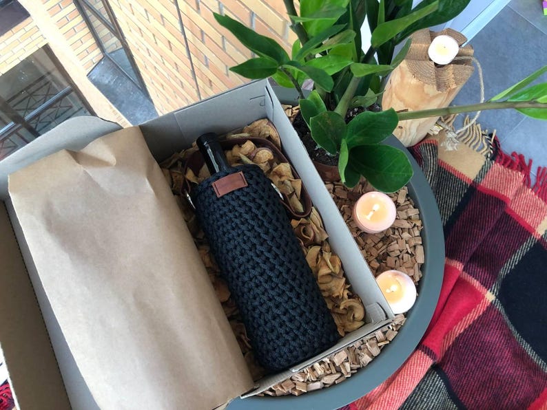 K&ouml;nnte beinhalten: Eine schwarze, gestrickte Weinflaschenh&uuml;lle mit einem braunen Lederetikett, pr&auml;sentiert in einem Karton mit braunem Papier und Holzsp&auml;nen. Die Box befindet sich in einem dekorativen Topf mit einer Pflanze, Kerzen und einer karierten Decke.