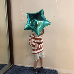 Peut inclure: Un ballon en forme d'&eacute;toile turquoise est tenu par une personne portant une chemise &agrave; manches longues ray&eacute;e et un short. Le ballon est brillant et refl&egrave;te la lumi&egrave;re. La personne se tient sur un tapis bleu.