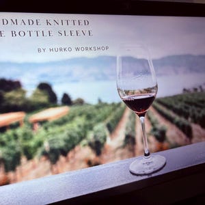 K&ouml;nnte beinhalten: Ein Weinglas mit Rotwein steht auf einer Fensterbank, im Hintergrund ein verschwommener Weinberg und See. Der Text oben lautet "HANDMADE KNITTED WINE BOTTLE SLEEVE" und "BY HURKO WORKSHOP".