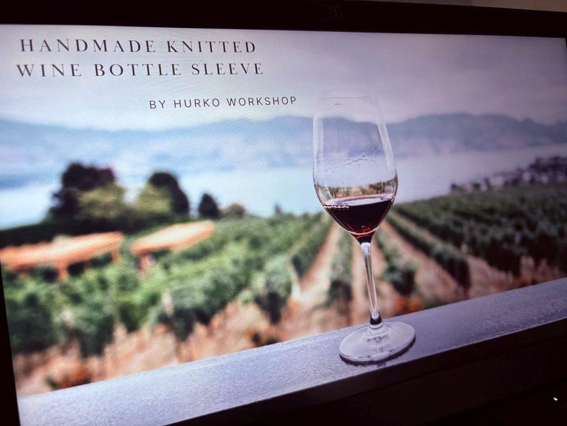 K&ouml;nnte beinhalten: Ein Weinglas mit Rotwein steht auf einer Fensterbank, im Hintergrund ein verschwommener Weinberg und See. Der Text oben lautet "HANDMADE KNITTED WINE BOTTLE SLEEVE" und "BY HURKO WORKSHOP".