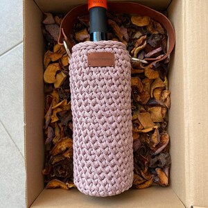 K&ouml;nnte beinhalten: Ein mauvefarbener, geh&auml;kelter Weinflaschenhalter mit einem braunen Lederriemen und einem kleinen, rechteckigen Lederetikett. Die Flasche befindet sich in einer Pappschachtel mit getrockneten Fr&uuml;chten. Der Flaschenhals ist sichtbar, mit dem Text "VELA UA".