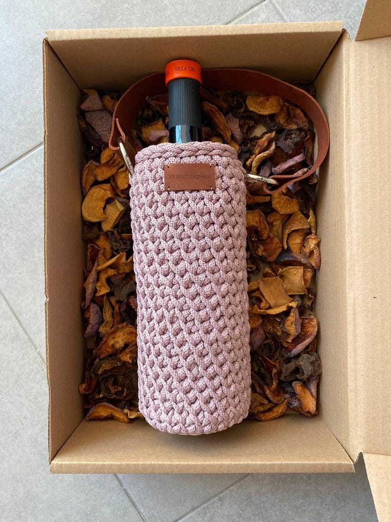 K&ouml;nnte beinhalten: Ein mauvefarbener, geh&auml;kelter Weinflaschenhalter mit einem braunen Lederriemen und einem kleinen, rechteckigen Lederetikett. Die Flasche befindet sich in einer Pappschachtel mit getrockneten Fr&uuml;chten. Der Flaschenhals ist sichtbar, mit dem Text "VELA UA".