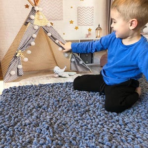 Peut inclure: Un tapis bleu textur&eacute; remplit le premier plan. Un enfant en chemise bleue et pantalon noir est assis pr&egrave;s d'un tipi de jeu. Le tipi est beige et gris avec des pompons blancs et une structure en bois. Des &eacute;toiles d&eacute;coratives et des motifs chevrons ornent le mur.