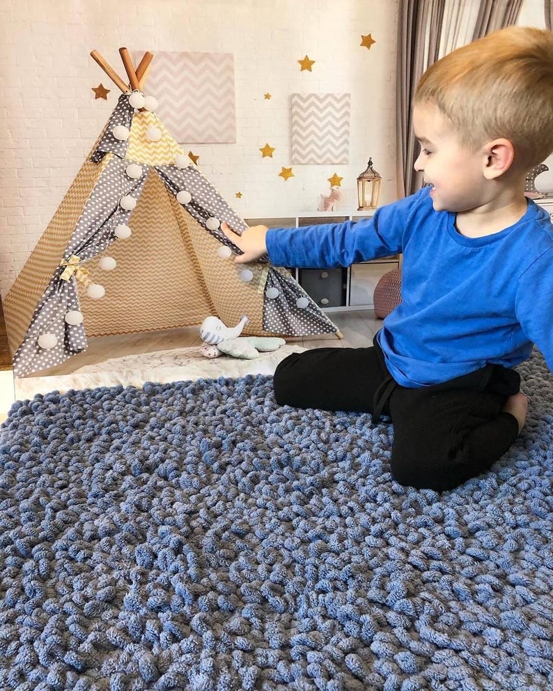 Peut inclure: Un tapis bleu textur&eacute; remplit le premier plan. Un enfant en chemise bleue et pantalon noir est assis pr&egrave;s d'un tipi de jeu. Le tipi est beige et gris avec des pompons blancs et une structure en bois. Des &eacute;toiles d&eacute;coratives et des motifs chevrons ornent le mur.