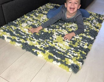 Puede incluir: Una alfombra rectangular hecha a mano en tonos grises, blancos y amarillos. La alfombra está hecha de hilo suave y en bucle y está colocada sobre un suelo de madera de color claro. La textura de la alfombra es visible y parece cómoda.