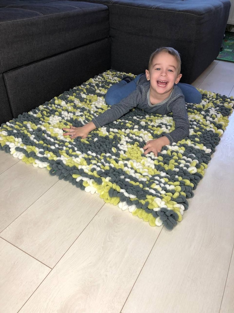 Peut inclure: Un tapis rectangulaire fait &agrave; la main avec un motif textur&eacute; en boucle. Le tapis pr&eacute;sente un m&eacute;lange de fils gris, blancs et vert clair. Le tapis est pos&eacute; sur un sol en bois de couleur claire.