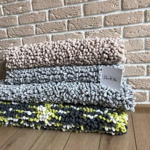 Peut inclure: Une pile de quatre couvertures en peluche textur&eacute;es de diff&eacute;rentes couleurs : beige, gris et un m&eacute;lange de gris, blanc et jaune. Les couvertures sont pli&eacute;es et empil&eacute;es les unes sur les autres. Une petite &eacute;tiquette blanche avec du texte est visible.