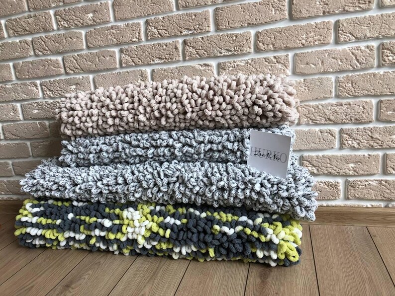 Peut inclure: Une pile de quatre couvertures en peluche textur&eacute;es de diff&eacute;rentes couleurs : beige, gris et un m&eacute;lange de gris, blanc et jaune. Les couvertures sont pli&eacute;es et empil&eacute;es les unes sur les autres. Une petite &eacute;tiquette blanche avec du texte est visible.