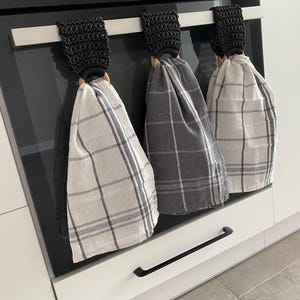 May include: Three kitchen towels hanging on a stainless steel bar. The towels have a plaid pattern in shades of gray and white. Each towel is held by a black crocheted loop and a wooden ring. The towels are hanging on a black oven.